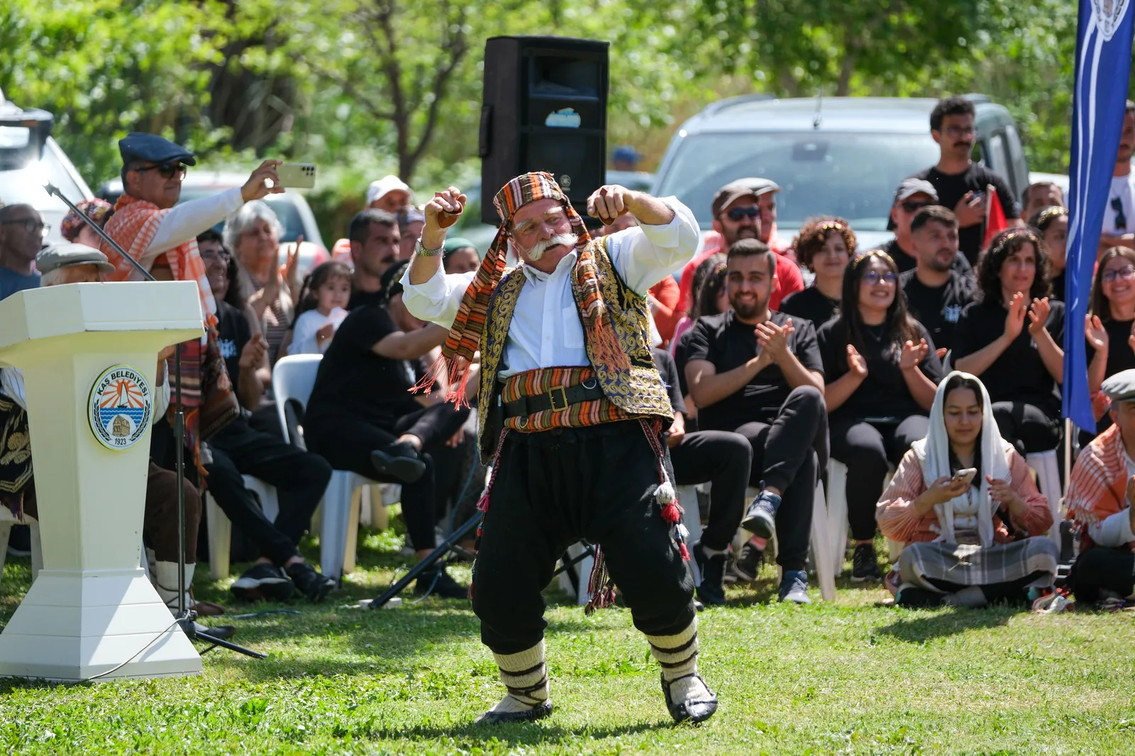 A man in a patterned vest, sash and baggy trousers dances in a park clearing in Antalya, with a seated audience clapping behind him. Not taken at this venue.