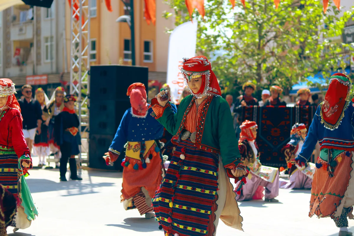 Dancers in striped, embroidered regional costume with red headscarves move in a line along a street at an outdoor festival, with a stage and onlookers behind