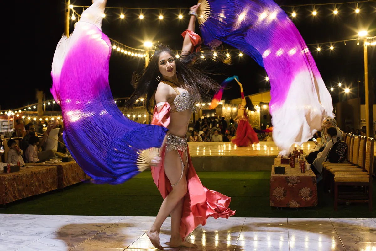 A belly dancer in a coral skirt and silver beaded top sweeps two large violet, magenta and white silk fans around her on a lit stage at night, with strings of lights above and diners at tables behind
