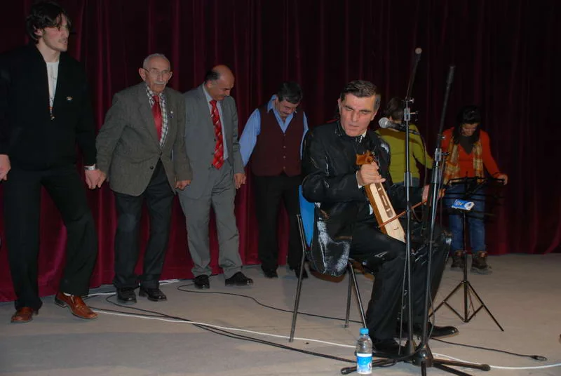 A line of men in jackets and ties dance with linked arms on a small stage while a seated musician plays a small fiddle